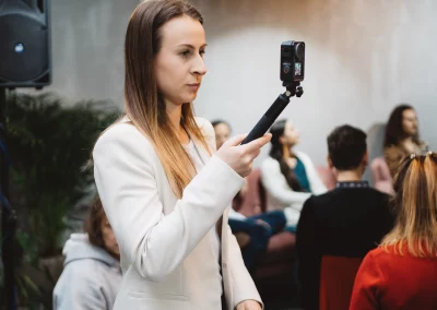 Eine Dame steht vor dem Hintergrund einer Podiumsdiskussion mit Zuschauern. Sie hält eine Kamera an einem Selfie-Stick und erfasst inspirierende Momente. Die Szene betont den Wunsch, Markenbotschafterin zu werden.