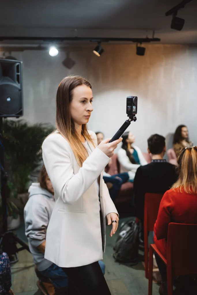 Markenbotschafter_werden Eine Dame steht vor dem Hintergrund einer Podiumsdiskussion mit Zuschauern. Sie hält eine Kamera an